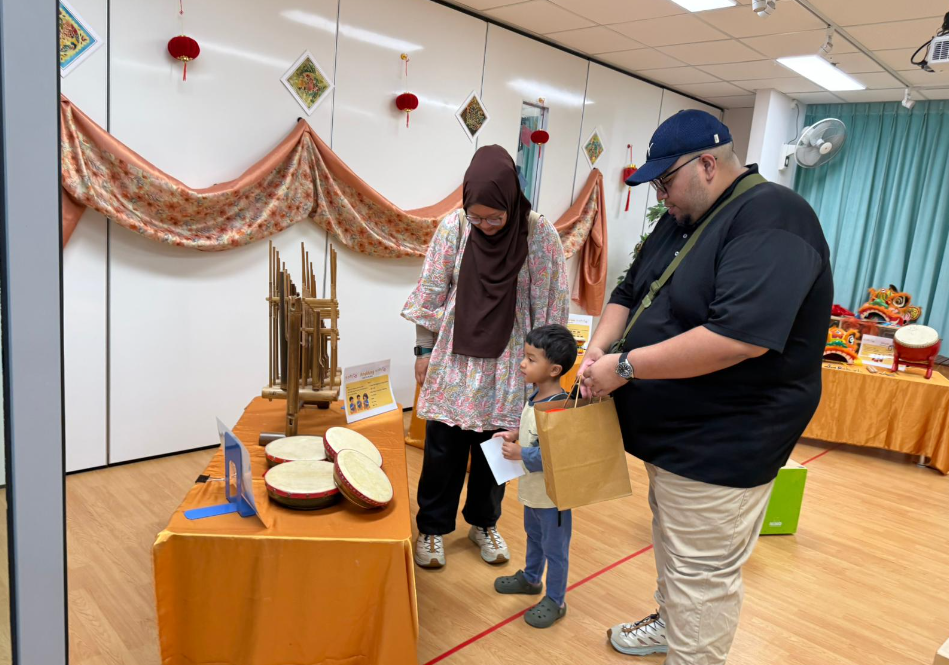 Parents and child look at drums and bamboo instruments on a table in a decorated indoor hall.
